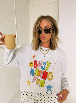 A woman wearing a light gray crewneck sweatshirt with a colorful 'BUSY HAVING FUN' graphic, styled with white polka dot pants, layered necklaces, and sunglasses, holding an iced coffee in a bright room. Find at Southern Quirks Gifts & Goods.