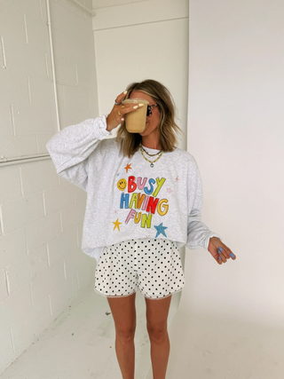 A woman takes a mirror selfie wearing a light gray heathered sweatshirt with a colorful 'BUSY HAVING FUN' graphic and white shorts with black polka dots. She is holding a coffee cup in a white-walled room.
