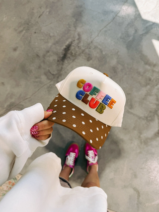 Person holding a polka dot hat with 'Coffee Club' text on it.  Standing on concrete floor.