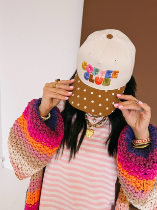 Person holding a cap with 'Coffee Club' text in front of their face, wearing a colorful sweater and striped dress.