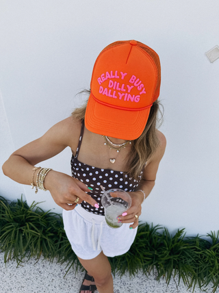 Girl holding a drink wearing an orange hat that says "really busy dilly dallying" in hot pink