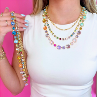 A woman wearing a white top models a multi-layered statement necklace with gold-tone chains and colorful rhinestones, holding up a matching strand of the necklace against a bright pink background.  Found at Southern Quirks Gifts & Goods in Edmond, Oklahoma.