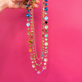 A hand holds up a long layered necklace with multiple gold-tone chains and colorful faceted rhinestone-like beads against a bright pink background. Taylor Shaye Designs
