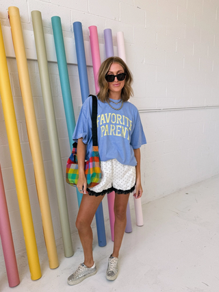 Woman in a blue oversized t-shirt with 'FAVORITE PARENT' text, holding a colorful bag, standing in front of tall, colorful cylindrical objects.