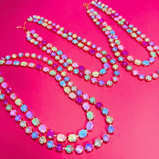 Three double-strand necklaces from Taylor Shaye with colorful iridescent rhinestones in pink, blue, green, and purple, set in gold-tone metal, arranged on a vibrant hot pink background. Found at Southern Quirks Gifts & Goods in Edmond, Oklahoma.