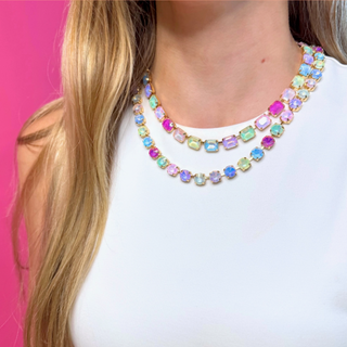 A close-up of a model wearing a double-strand gold necklace with colorful rectangular iridescent gemstones against a bright pink background. Found at southern quirks gifts & goods in Edmond, OK.