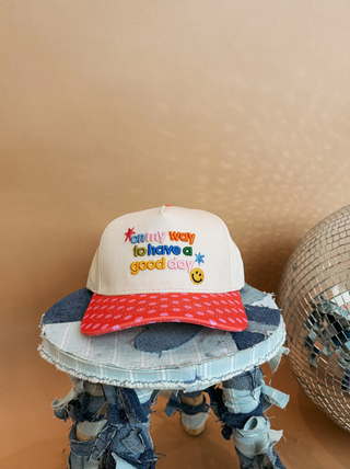 A white baseball cap with a red brim sits on a distressed denim stool. The cap features colorful embroidered text reading 'creative way to have a good day' with a smiley face and stars.  Colorful, quirky, cheerful hat from Southern Quirks Gifts & Goods.