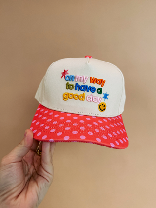 A hand holds a white and pink trucker-style cap with colorful embroidered text reading 'on my way to have a good day' and a smiley face on the front, against a plain beige background.  Shop at Southern Quirks Gifts & Goods.