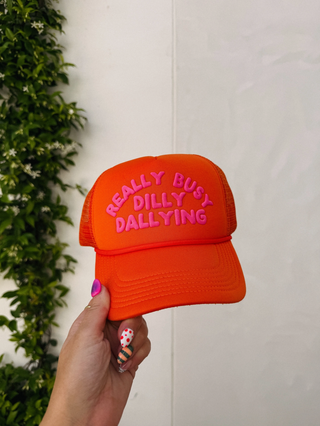 Orange cap with pink text held by a hand against a white wall with greenery.