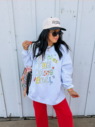 A woman stands against a white wooden wall wearing a light blue heathered sweatshirt with colorful text reading 'RUN THE DANCE BALL,' red pants, a white cap, sunglasses, and a patterned shoulder bag.  Etta + East label found at Southern Quirks Gifts & Goods