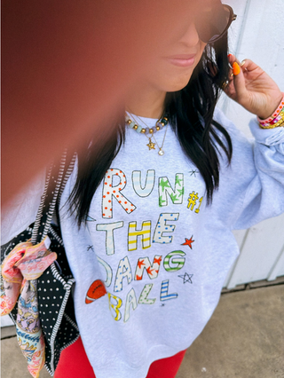 A person wearing a light blue graphic sweatshirt with colorful retro-style text reading 'RUN THE DANG BALL!' and small space-themed doodles, paired with red pants and layered gold necklaces, photographed outdoors against a light wall.  Cute and quirky style for moms and football fans.
