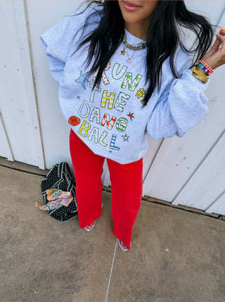 A woman wearing a light blue graphic sweatshirt with colorful text reading 'RUN THE DANG BALL' and red pants, standing against a white paneled wall.