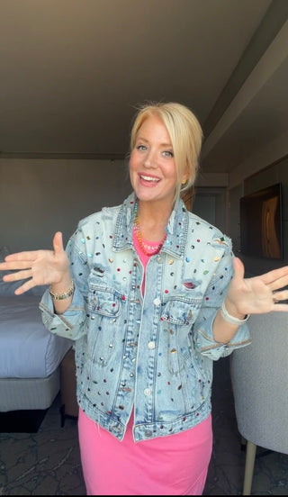 A smiling blonde woman stands in a hotel room wearing a light-wash denim jacket covered in small colorful embroidered patches, layered over a bright pink top and pink skirt. She has her hands out in a welcoming gesture.