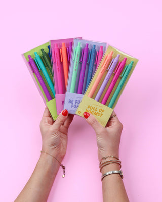 Four packs of pastel-colored gel pens held up by two hands against a solid pink background. The pens are in clear plastic packaging with colorful paper sleeves showing shades of green, blue, purple, pink, orange, and yellow. Located at Southern Quirks Gifts & Goods