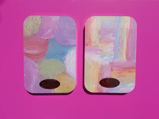 Two rectangular wall art panels with rounded corners, featuring pastel abstract painted designs in pink, purple, blue, yellow, and cream, mounted against a bright pink background. Each panel has a small oval dark plaque near the bottom.