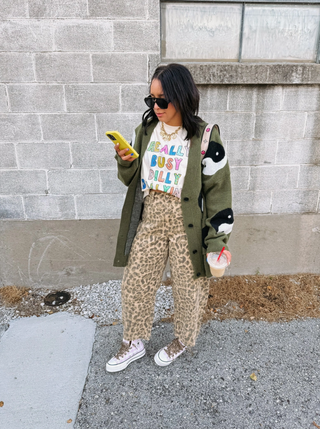 Person wearing a green cardigan with animal print patterns, standing against a gray brick wall.
