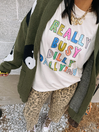 Person wearing a colorful 'REALLY BUSY DILLY DALLYING' shirt with a green cardigan and leopard print pants.